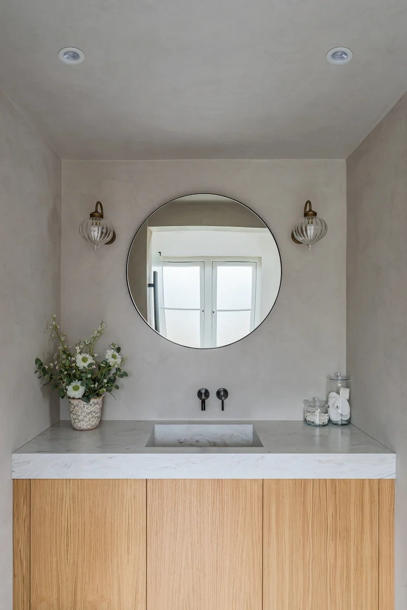 Warm greige taupe microcement powder room in Park City, Utah with round black mirror, ribbed glass, brass sconces, marble trough basin and oak vanity