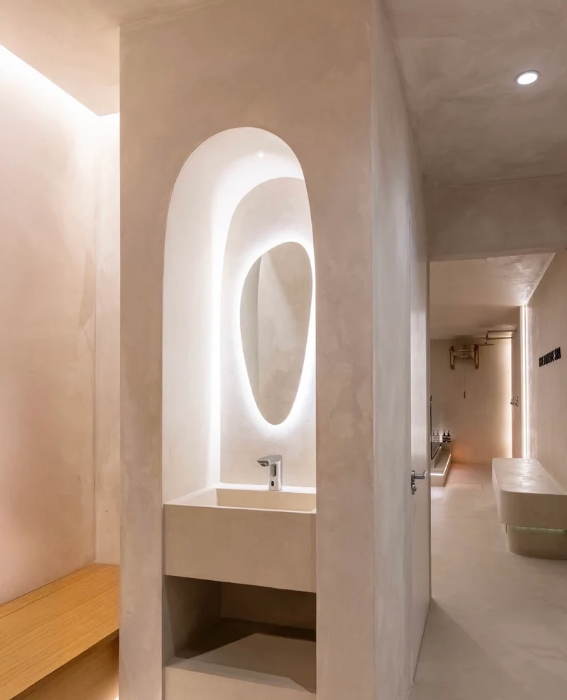 Warm blush oat microcement washroom in Park City, Utah with arched niche, backlit organic oval mirror, cast basin and trough vanity