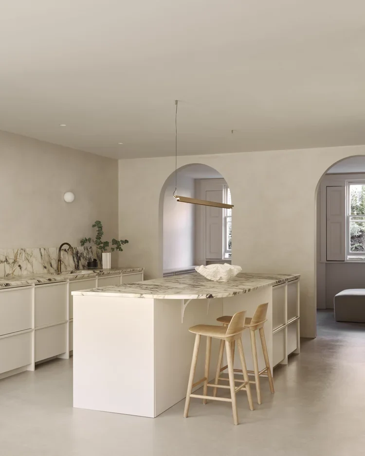 Bone oatmeal microcement kitchen walls and greige floor in Park City, Utah with arched openings, violet-veined marble island, cream cabinetry and oak stools