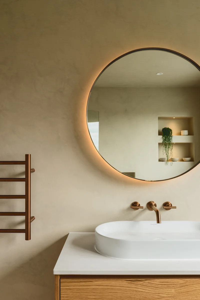 Warm greige microcement vanity wall in Park City, Utah with backlit round mirror, brushed copper tap and white oak floating vanity