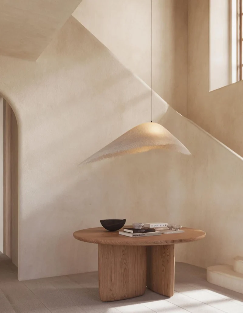 Warm ecru blush microcement stair hall walls in Park City, Utah with sculptural soffit, diagonal sunlight shaft, woven pendant and round oak table